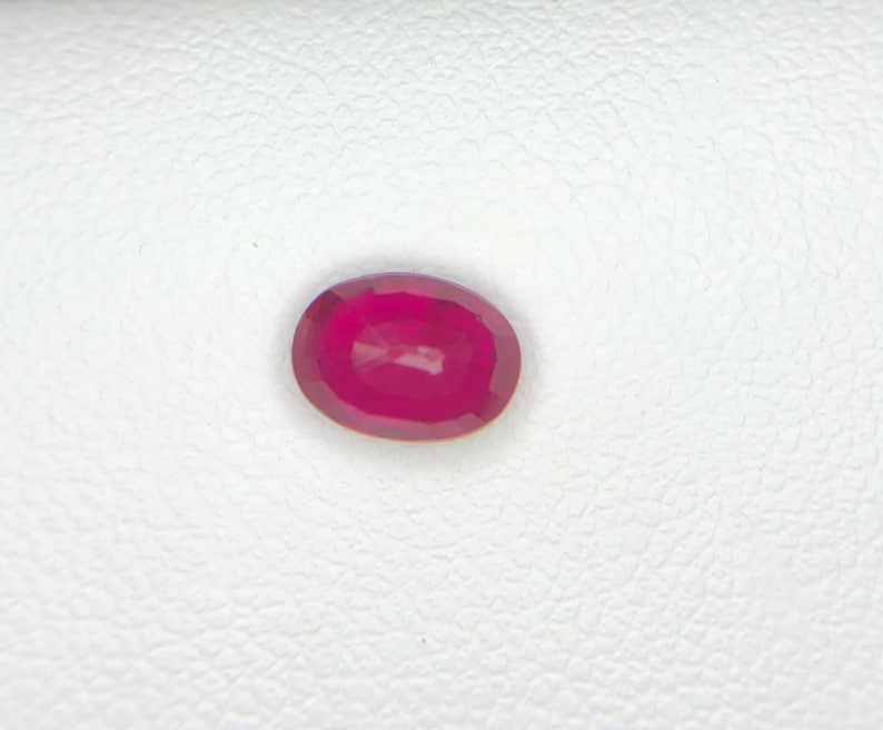 Peut inclure: Une pierre pr&eacute;cieuse taill&eacute;e en ovale, d'une couleur rose-rouge profonde et vibrante. La pierre pr&eacute;sente une surface &agrave; facettes qui refl&egrave;te la lumi&egrave;re, mettant en valeur sa qualit&eacute; translucide et ses bords arrondis sur un fond blanc textur&eacute;.