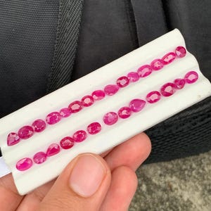 May include: A collection of vibrant, faceted, deep pinkish-red gemstones arranged in two rows on a white display tray. The stones feature various shapes, including oval, pear, and round cuts, all exhibiting a polished, reflective surface.