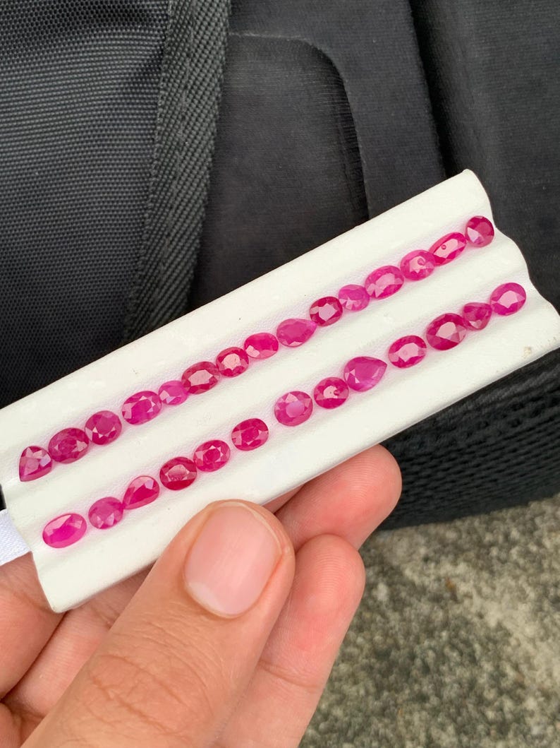 May include: A collection of vibrant, faceted, deep pinkish-red gemstones arranged in two rows on a white display tray. The stones feature various shapes, including oval, pear, and round cuts, all exhibiting a polished, reflective surface.