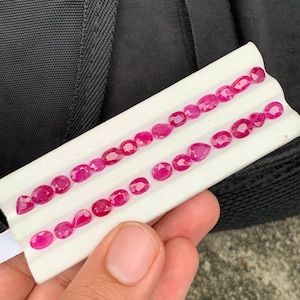 May include: A collection of small, faceted, vibrant pinkish-red gemstones arranged in two rows on a white display card. The stones vary in shape, including oval, pear, and round cuts, each exhibiting a polished surface that reflects light.