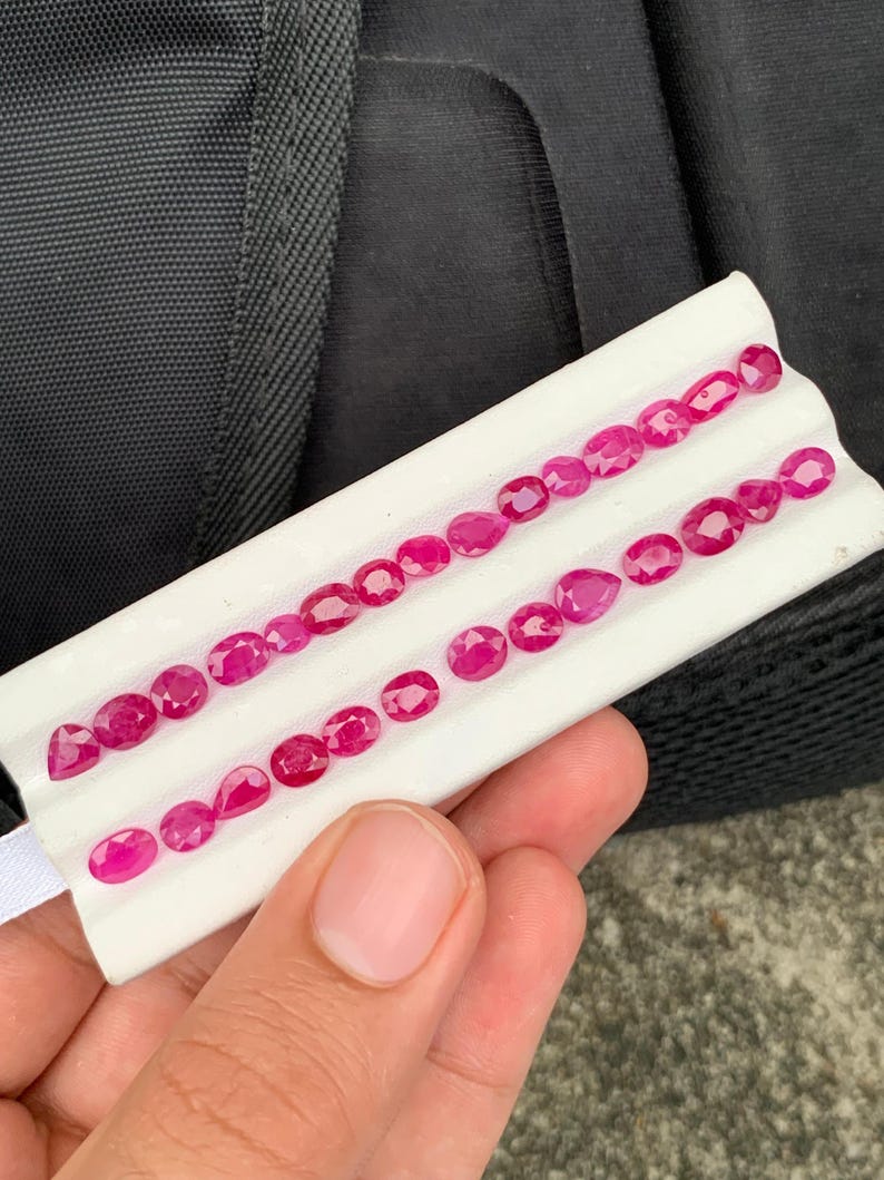 May include: A collection of small, faceted, vibrant pinkish-red gemstones arranged in two rows on a white display card. The stones vary in shape, including oval, pear, and round cuts, each exhibiting a polished surface that reflects light.