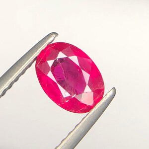 Peut inclure: Une pierre pr&eacute;cieuse rubis de taille ovale, d'une teinte rouge ros&eacute;e vive et dot&eacute;e d'une surface &agrave; facettes, maintenue fermement entre les deux pointes m&eacute;talliques d'une pince, r&eacute;v&eacute;lant sa clart&eacute; interne et ses reflets lumineux.