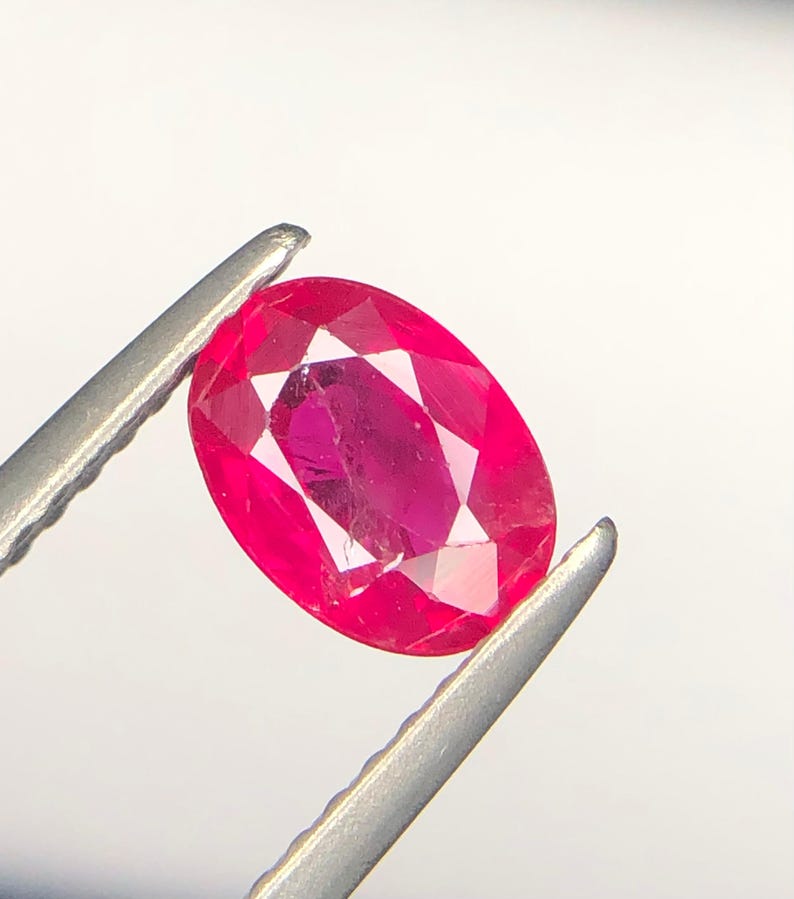 Peut inclure: Une pierre pr&eacute;cieuse rubis de taille ovale, d'une teinte rouge ros&eacute;e vive et dot&eacute;e d'une surface &agrave; facettes, maintenue fermement entre les deux pointes m&eacute;talliques d'une pince, r&eacute;v&eacute;lant sa clart&eacute; interne et ses reflets lumineux.
