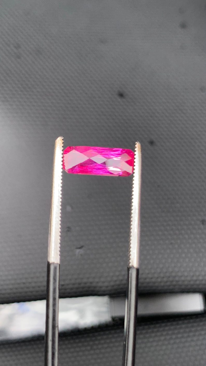 May include: A rectangular, faceted gemstone with a vibrant, saturated hot pink colour. The stone is held horizontally by a pair of metal tweezers with serrated tips, showcasing its clear, polished surface and geometric cut against a dark background.