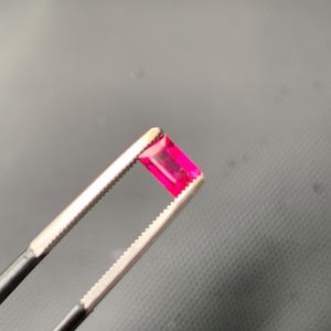 May include: A vibrant, rectangular-cut gemstone with a deep pinkish-red hue held securely between the serrated metal tips of a pair of professional jeweler's tweezers. The faceted stone displays clear light reflection and a polished surface.