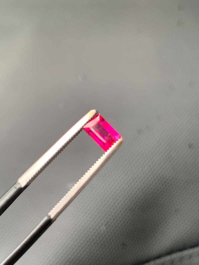 May include: A vibrant, rectangular-cut gemstone with a deep pinkish-red hue held securely between the serrated metal tips of a pair of professional jeweler's tweezers. The faceted stone displays clear light reflection and a polished surface.