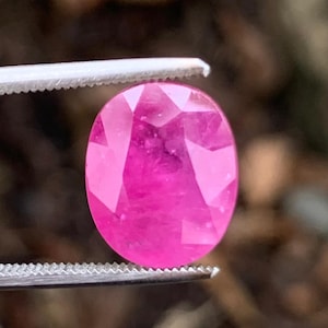以下が含まれることがあります： 金属製のピンセットで保持された、ファセットカットが施された鮮やかなピンク色の楕円形の宝石。この石は半透明で内部にインクルージョンが見られ、磨かれた構造全体に明るく彩度の高いマゼンタピンクの色合いが広がっています。