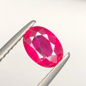 Pode incluir: Uma gema de corte oval com um tom rosa-avermelhado profundo e saturado. A pedra apresenta uma superfície facetada que reflete a luz, exibindo a sua clareza e estrutura interna. É mantida firmemente por uma pinça metálica de tom prateado.