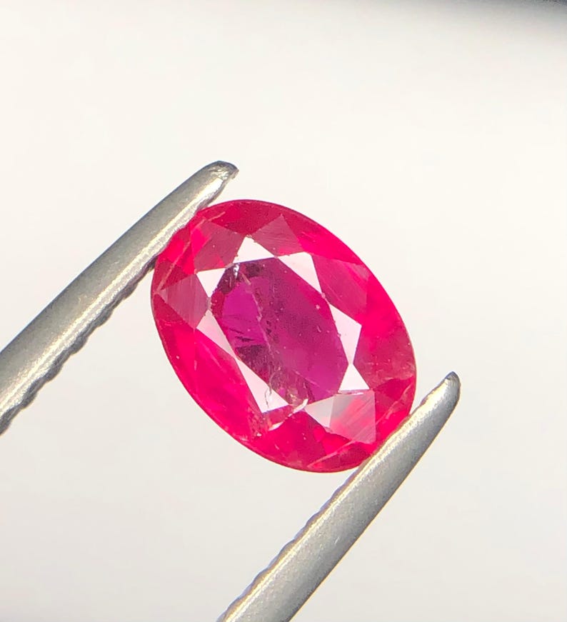 Peut inclure: Une pierre pr&eacute;cieuse de taille ovale d'une teinte rose-rouge profonde et satur&eacute;e. La pierre pr&eacute;sente une surface &agrave; facettes qui refl&egrave;te la lumi&egrave;re, mettant en valeur sa clart&eacute; et sa structure interne. Elle est tenue par une pince m&eacute;tallique argent&eacute;e.
