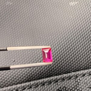 May include: A vibrant, rectangular-cut pink gemstone held by a pair of metal tweezers. The stone features a faceted surface that reflects light, showcasing a deep, saturated magenta hue with clear, sharp edges and a polished, transparent appearance.