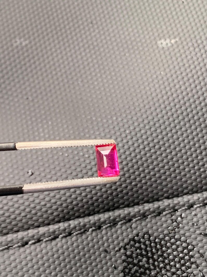 May include: A vibrant, rectangular-cut pink gemstone held by a pair of metal tweezers. The stone features a faceted surface that reflects light, showcasing a deep, saturated magenta hue with clear, sharp edges and a polished, transparent appearance.