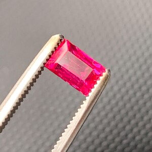 May include: A vibrant, rectangular-cut gemstone with a deep, saturated pinkish-red hue. The stone features a faceted surface that reflects light, showcasing its clarity and color. It is held securely by a pair of metallic, serrated-edge tweezers.