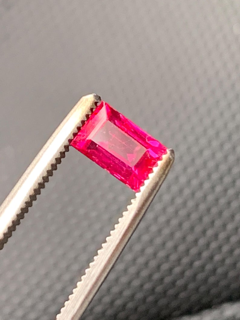 May include: A vibrant, rectangular-cut gemstone with a deep, saturated pinkish-red hue. The stone features a faceted surface that reflects light, showcasing its clarity and color. It is held securely by a pair of metallic, serrated-edge tweezers.