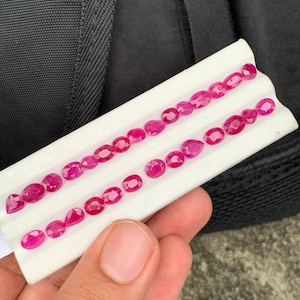 May include: A collection of small, faceted, vibrant pinkish-red gemstones arranged in two rows on a white display card. The stones feature various cuts, including oval, pear, and round shapes, showcasing a brilliant, reflective surface texture.