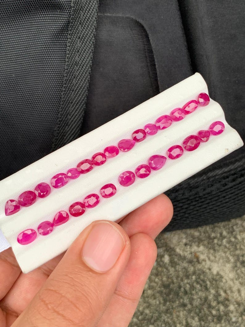 May include: A collection of small, faceted, vibrant pinkish-red gemstones arranged in two rows on a white display card. The stones feature various cuts, including oval, pear, and round shapes, showcasing a brilliant, reflective surface texture.