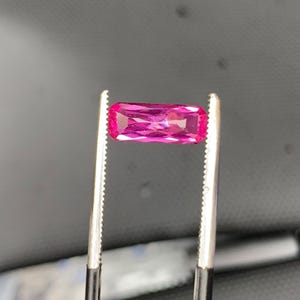 May include: A vibrant, hot pink gemstone with a rectangular baguette cut, featuring multiple facets that reflect light. The gemstone is held securely by a pair of metal tweezers with serrated tips, showcasing its clarity and deep, saturated colour.