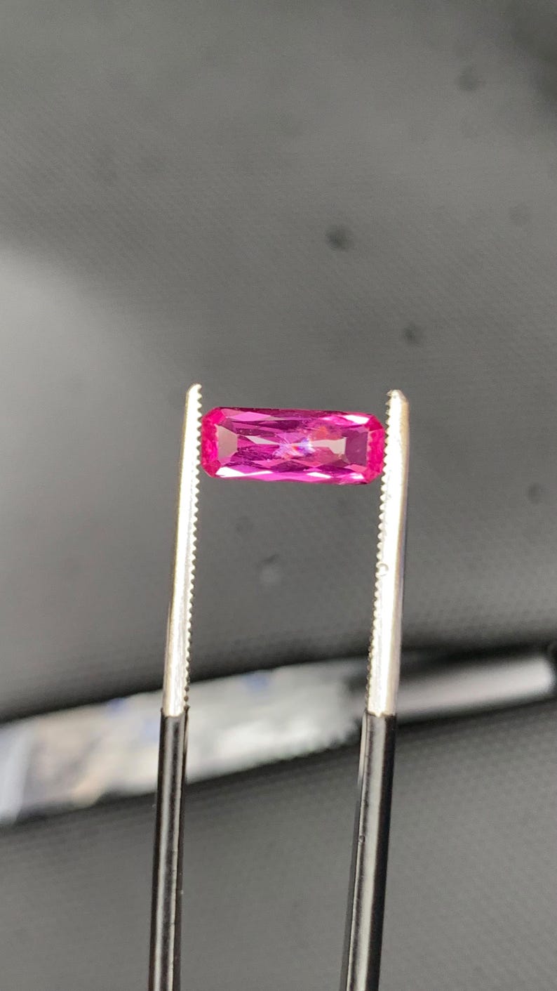 May include: A vibrant, hot pink gemstone with a rectangular baguette cut, featuring multiple facets that reflect light. The gemstone is held securely by a pair of metal tweezers with serrated tips, showcasing its clarity and deep, saturated colour.