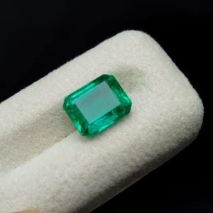 Può includere: Uno smeraldo dal taglio rettangolare con una tonalità verde intenso e superfici sfaccettate. La pietra preziosa è inserita in un morbido cuscinetto da esposizione testurizzato color bianco sporco, che ne esalta la chiarezza e la luce.