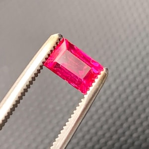 May include: A vibrant, rectangular-cut gemstone with a deep pinkish-red hue, held securely by a pair of metallic tweezers. The stone features a faceted surface that reflects light, showcasing its clear, transparent quality and sharp, defined edges.