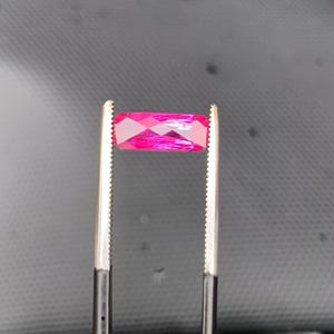 May include: A vibrant, rectangular, faceted gemstone in a deep magenta-pink hue, held securely by a pair of metal tweezers with serrated tips. The stone exhibits a clear, polished surface with multiple geometric facets that reflect light.