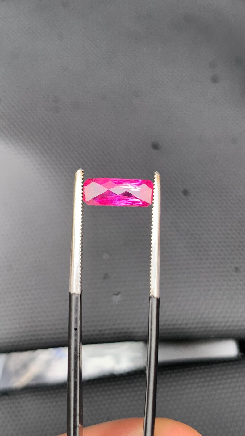 May include: A vibrant, rectangular, faceted gemstone in a deep magenta-pink hue, held securely by a pair of metal tweezers with serrated tips. The stone exhibits a clear, polished surface with multiple geometric facets that reflect light.
