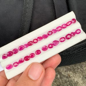 May include: A collection of small, faceted, vibrant red rubies arranged in two rows on a white display tray. The gemstones feature various shapes, including oval, round, and pear cuts, showcasing a deep crimson hue and reflective surfaces.