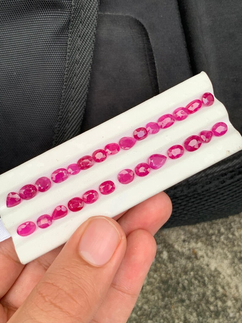 May include: A collection of small, faceted, vibrant red rubies arranged in two rows on a white display tray. The gemstones feature various shapes, including oval, round, and pear cuts, showcasing a deep crimson hue and reflective surfaces.