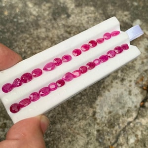 May include: A collection of vibrant, faceted, deep pink gemstones arranged in two parallel rows on a white display card. The stones are primarily oval and pear-shaped, exhibiting a brilliant cut that reflects light across their polished surfaces.