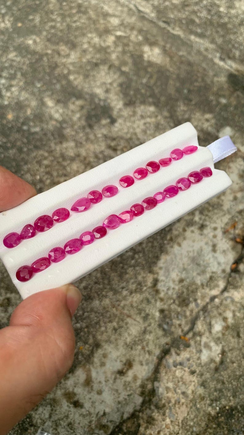 May include: A collection of vibrant, faceted, deep pink gemstones arranged in two parallel rows on a white display card. The stones are primarily oval and pear-shaped, exhibiting a brilliant cut that reflects light across their polished surfaces.