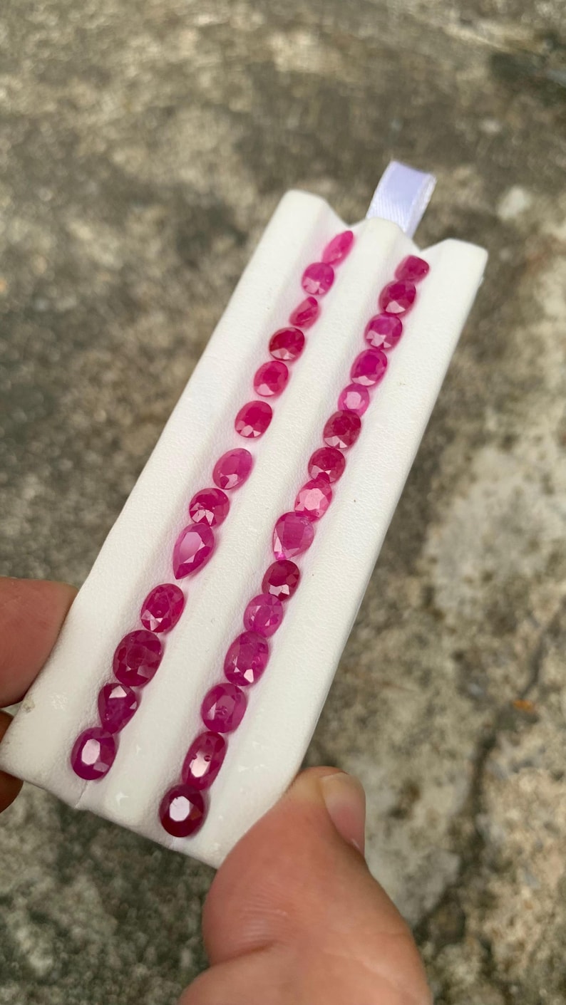 May include: A collection of vibrant, faceted, oval-shaped rubies arranged in two parallel rows on a white foam display holder. The gemstones exhibit a deep, saturated pinkish-red hue and are polished to reflect light across their multiple surfaces.