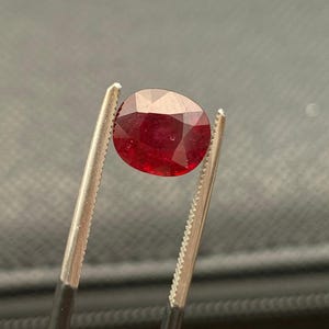 May include: A deep red, oval-cut gemstone held by a pair of metal tweezers. The stone features a faceted surface that reflects light, showcasing a rich, saturated crimson hue throughout its transparent body, captured in a clear, focused view.