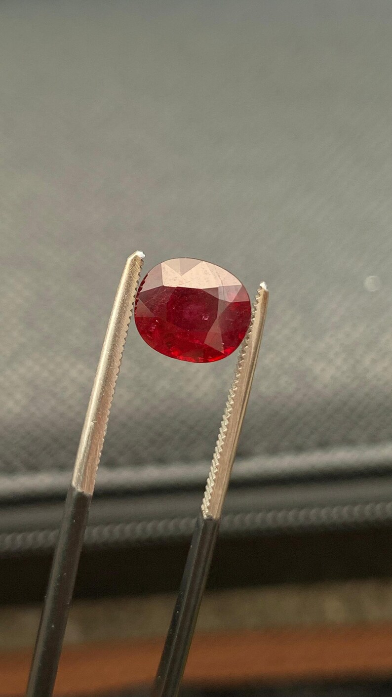 May include: A deep red, oval-cut gemstone held by a pair of metal tweezers. The stone features a faceted surface that reflects light, showcasing a rich, saturated crimson hue throughout its transparent body, captured in a clear, focused view.