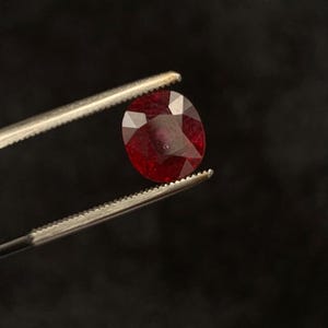 May include: A faceted, oval-shaped deep red gemstone held by a pair of metal tweezers. The stone features a polished surface with multiple geometric facets that reflect light, showcasing a rich, saturated crimson colour throughout the entire gem.