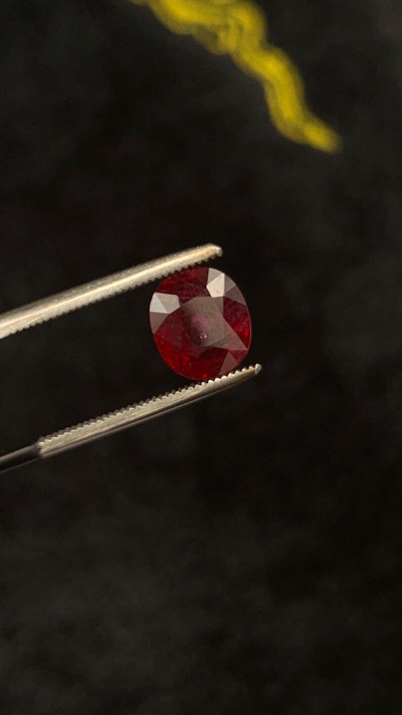 May include: A faceted, oval-shaped deep red gemstone held by a pair of metal tweezers. The stone features a polished surface with multiple geometric facets that reflect light, showcasing a rich, saturated crimson colour throughout the entire gem.