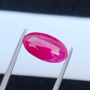 May include: A vibrant, oval-shaped, cabochon-cut gemstone in a deep, saturated magenta-pink color. The stone features a smooth, polished, and reflective surface that catches the light, showcasing its translucent quality and rich, uniform hue.