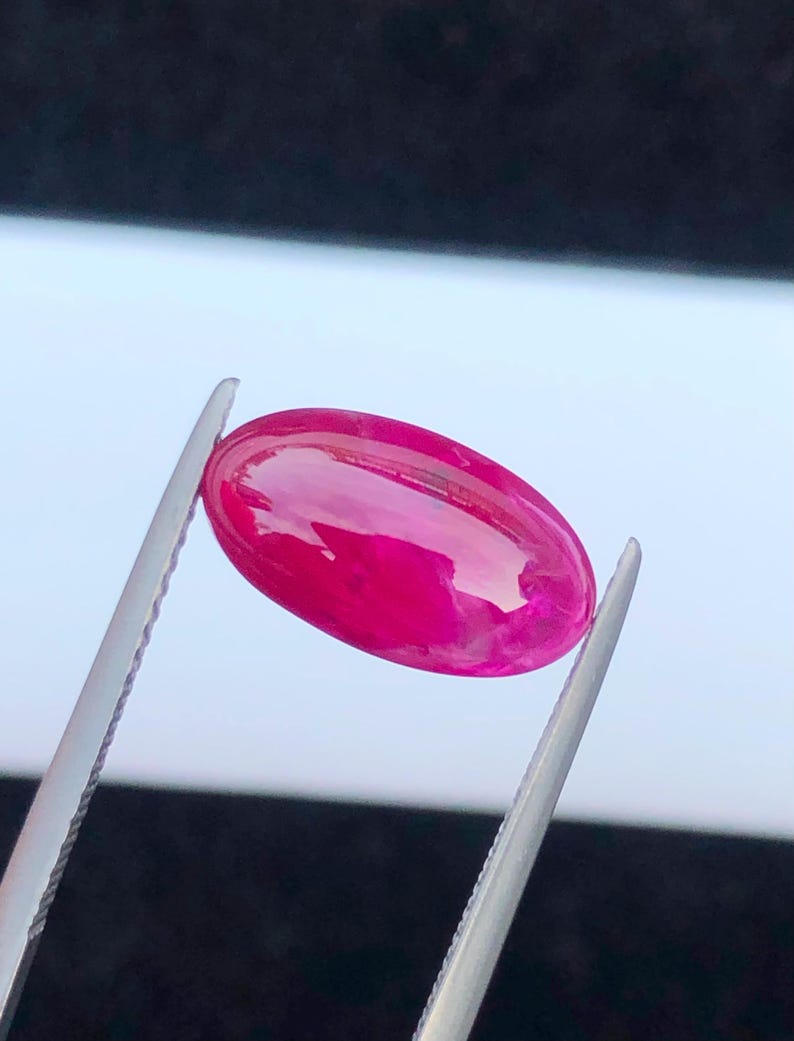 May include: A vibrant, oval-shaped, cabochon-cut gemstone in a deep, saturated magenta-pink color. The stone features a smooth, polished, and reflective surface that catches the light, showcasing its translucent quality and rich, uniform hue.