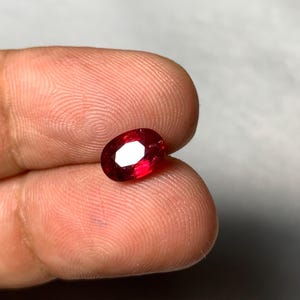 Op de afbeelding: Een levendige, ovaal geslepen edelsteen met een diepe, verzadigde rode kleur. De steen heeft meerdere facetten die het licht reflecteren, waardoor een schitterende glans over het oppervlak ontstaat. De edelsteen wordt tussen twee vingers gehouden.