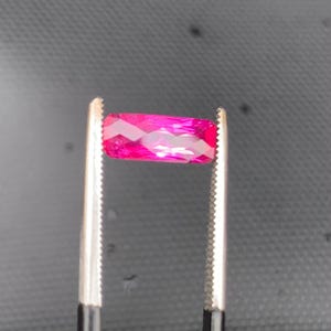 May include: A vibrant, rectangular-cut gemstone with a deep magenta-pink hue and multifaceted surface, held securely between the serrated metal tips of a pair of tweezers. The stone displays a brilliant, reflective quality with sharp, geometric edges.