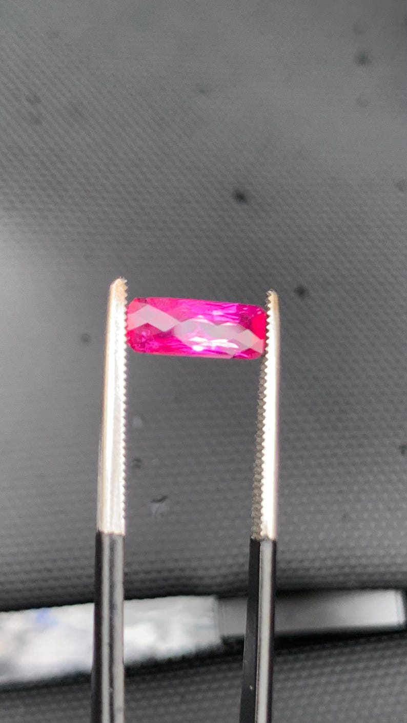 May include: A vibrant, rectangular-cut gemstone with a deep magenta-pink hue and multifaceted surface, held securely between the serrated metal tips of a pair of tweezers. The stone displays a brilliant, reflective quality with sharp, geometric edges.