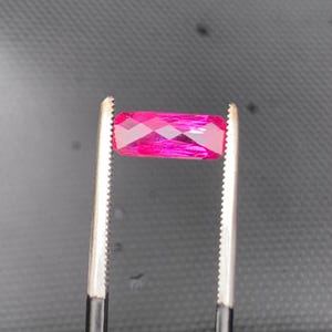 May include: A vibrant, rectangular, faceted gemstone in a deep magenta-pink hue. The stone is held horizontally by a pair of metal tweezers with serrated tips, showcasing its clear, polished surfaces and geometric cut against a dark background.