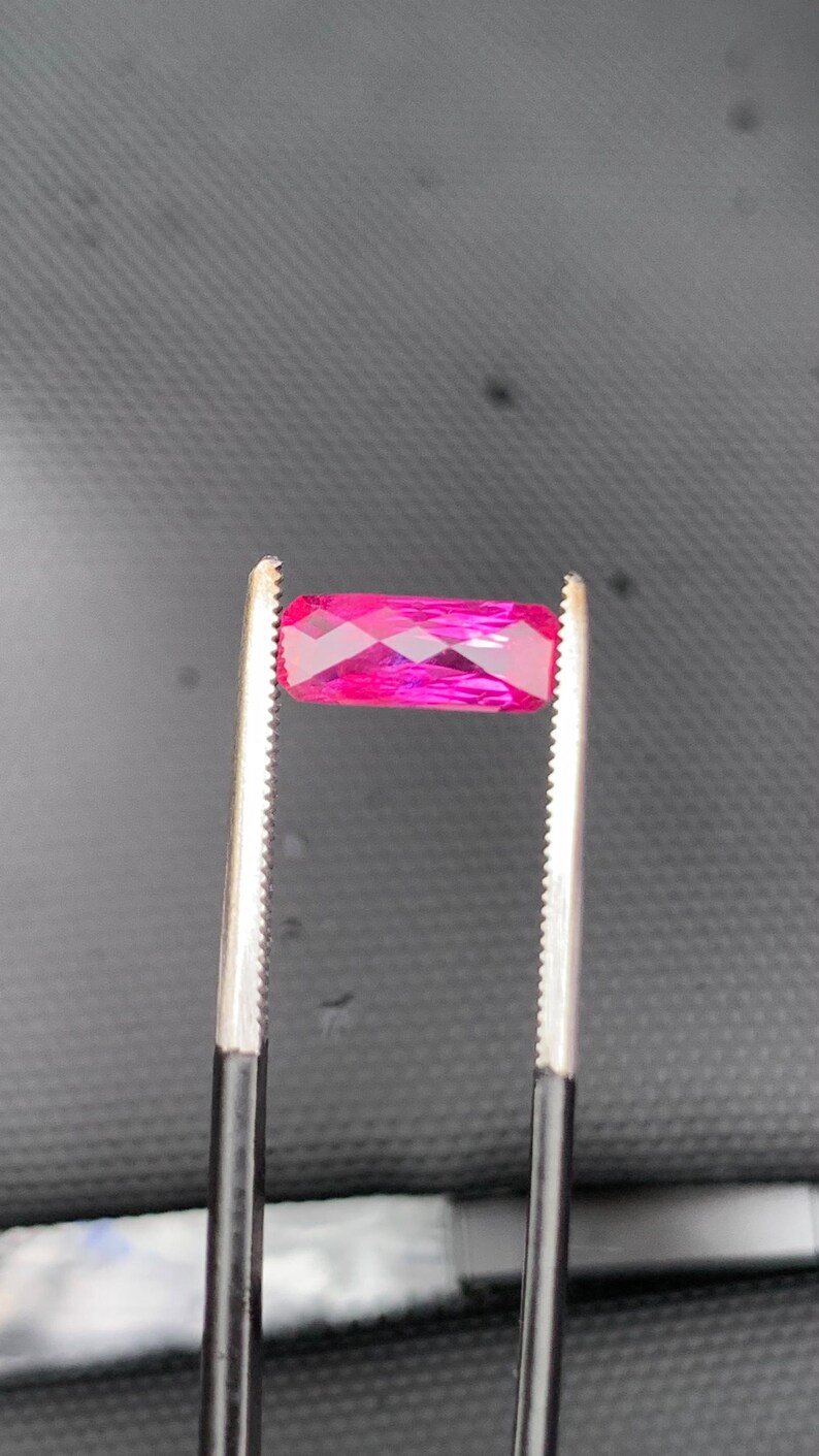 May include: A vibrant, rectangular, faceted gemstone in a deep magenta-pink hue. The stone is held horizontally by a pair of metal tweezers with serrated tips, showcasing its clear, polished surfaces and geometric cut against a dark background.