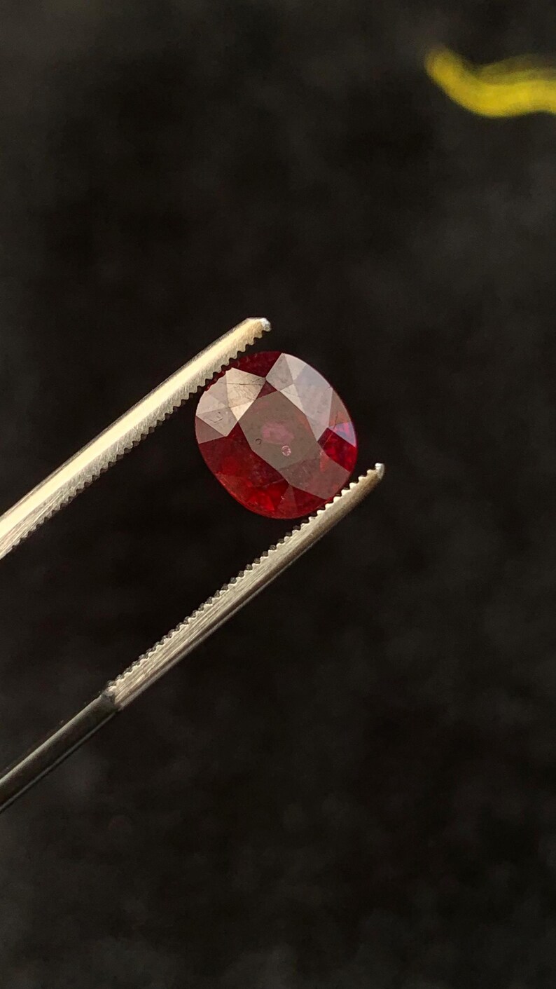 May include: A faceted, oval-shaped deep red gemstone held by a pair of silver-toned metal tweezers. The stone displays a translucent quality with visible internal inclusions and a polished surface that reflects light across its multiple cut facets.