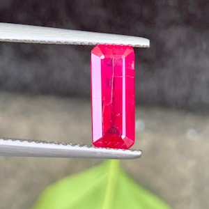 Puede incluir: Una gema de rubí de corte rectangular con un tono rojo intenso e inclusiones internas visibles. La gema está sujeta por unas pinzas de metal, mostrando su superficie facetada y sus bordes claros y pulidos.
