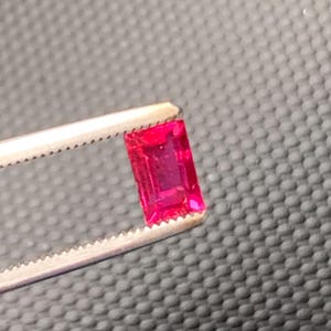 May include: A vibrant, rectangular, emerald-cut gemstone with a deep, saturated red color. The gemstone is held securely by a pair of silver-toned metal tweezers with serrated tips, showcasing the faceted surface and clarity of the red stone.