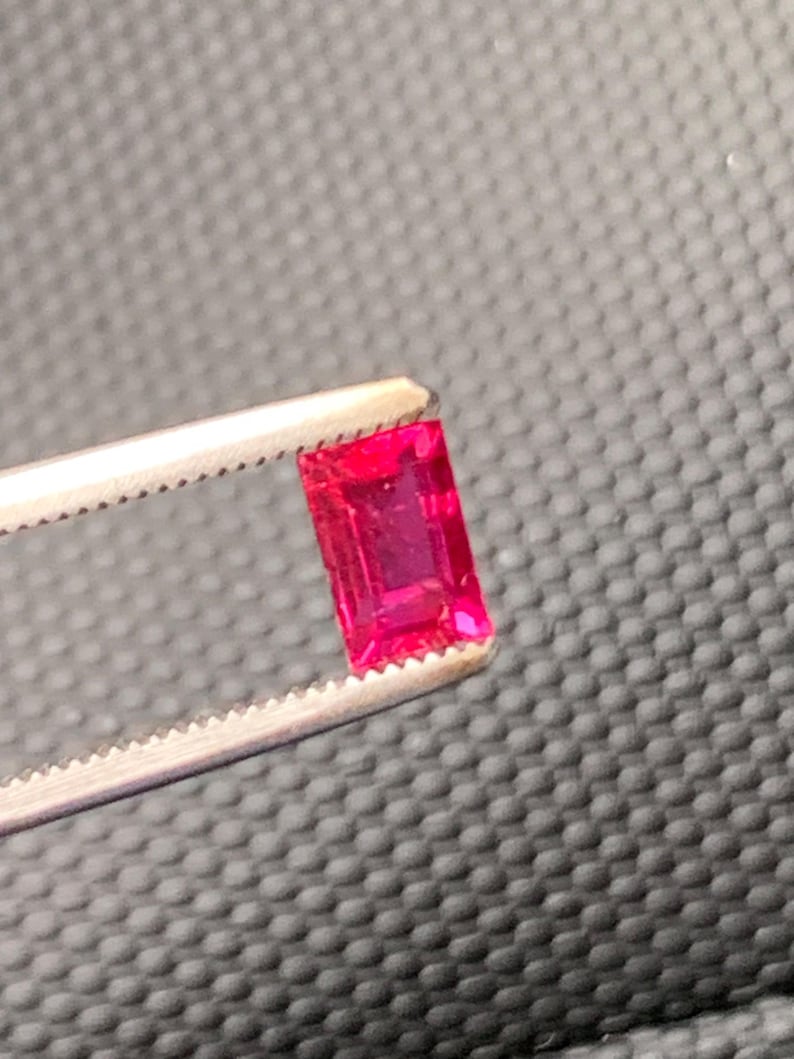 May include: A vibrant, rectangular, emerald-cut gemstone with a deep, saturated red color. The gemstone is held securely by a pair of silver-toned metal tweezers with serrated tips, showcasing the faceted surface and clarity of the red stone.