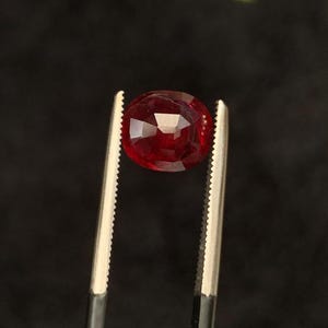 May include: A deep red, oval-cut gemstone held by a pair of silver-toned metal tweezers. The stone features multiple facets that reflect light, creating a brilliant appearance. The gemstone is positioned centrally, showcasing its rich, saturated colour.