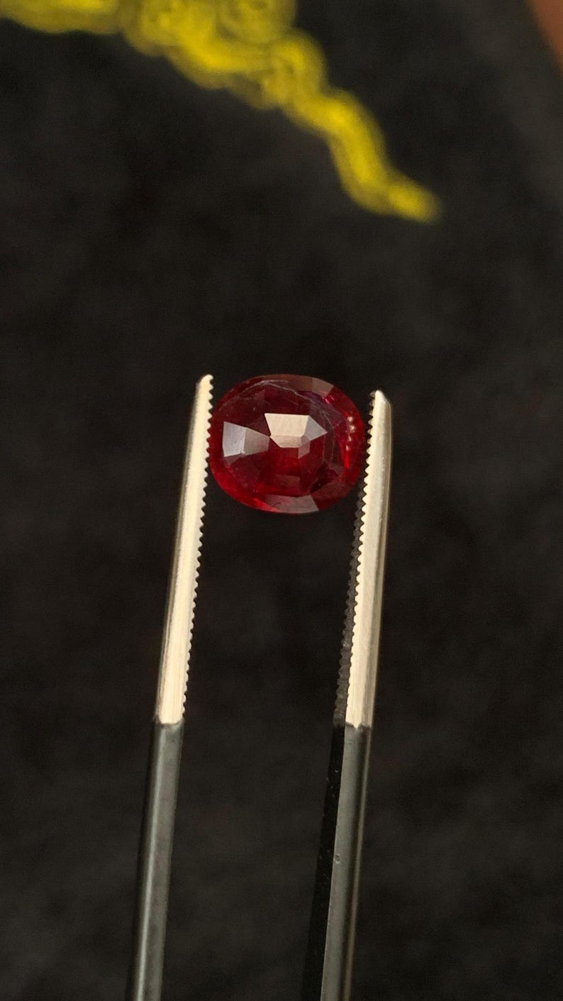 May include: A deep red, oval-cut gemstone held by a pair of silver-toned metal tweezers. The stone features multiple facets that reflect light, creating a brilliant appearance. The gemstone is positioned centrally, showcasing its rich, saturated colour.