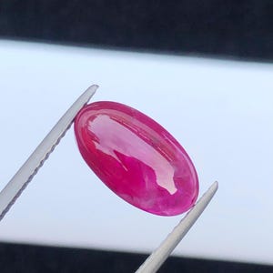 May include: A vibrant, oval-shaped, cabochon-cut gemstone with a deep magenta-pink hue and a smooth, polished surface. The stone is held by a pair of silver-toned metal tweezers, showcasing its translucent quality and reflective, glossy finish.
