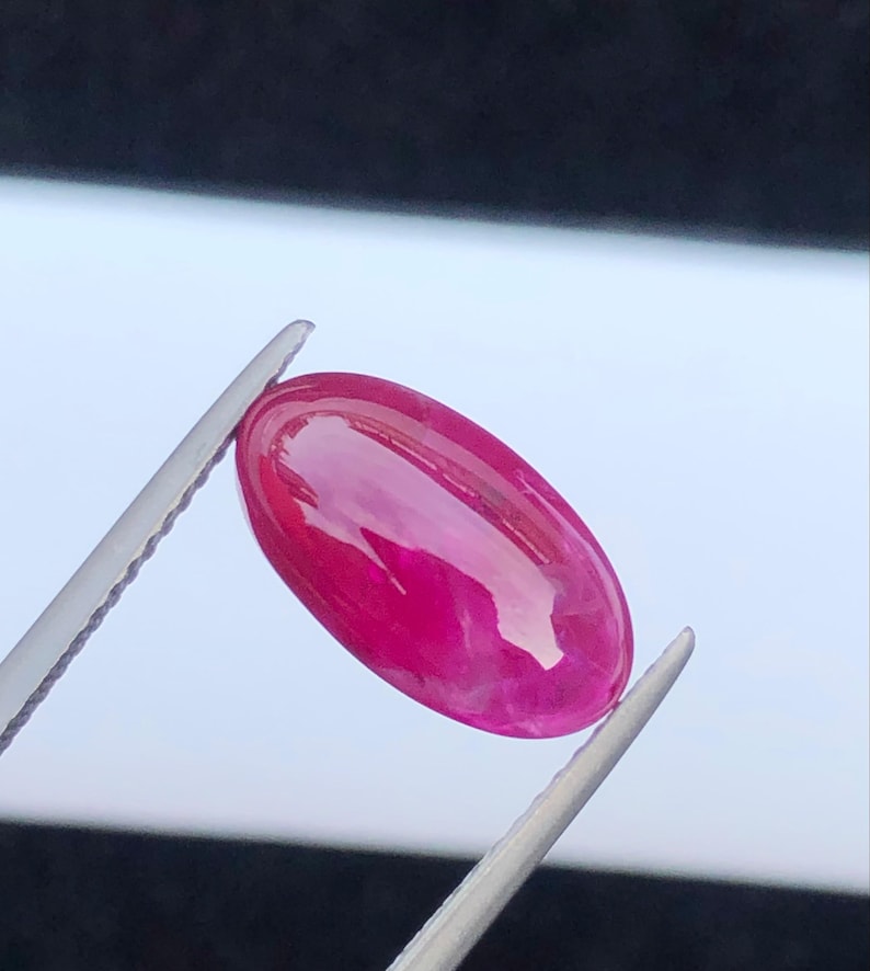 May include: A vibrant, oval-shaped, cabochon-cut gemstone with a deep magenta-pink hue and a smooth, polished surface. The stone is held by a pair of silver-toned metal tweezers, showcasing its translucent quality and reflective, glossy finish.