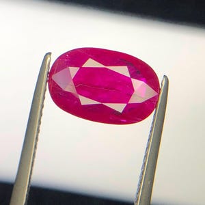 Puede incluir: Una piedra preciosa de corte ovalado en color rojo magenta vibrante sostenida por unas pinzas de metal plateado. La piedra presenta una superficie facetada con un patrón en forma de estrella que refleja la luz, mostrando una calidad translúcida.